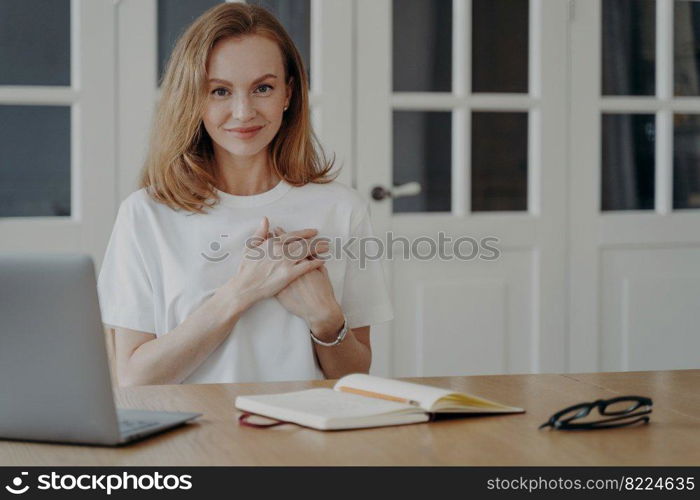 Woman folds hands close to heart and loves her remote job. Successful businesswoman working from home. Lovely european lady is sitting at desk in apartment. Concept of love and emotional expression.. Woman folds hands close to heart and loves her remote job. Successful businesswoman works from home.