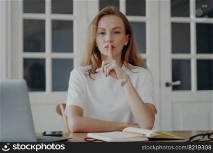Woman gestures hush. Successful mid adult businesswoman is working from home. Blond european lady in white t-shirt is an assistant having remote work in her apartment. Concept of secret and privacy.. Successful mid adult businesswoman is working from home and gestures hush. Concept of secret.