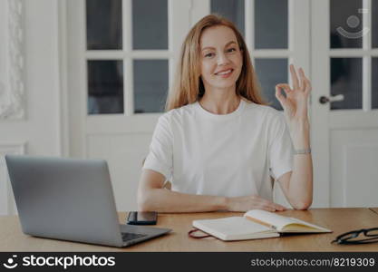 Woman gestures ok sitting at the desk in her apartment. Successful mid adult businesswoman. Lovely european lady in white t-shirt is freelancer, entrepreneur or executive having remote work.. Woman gestures ok sitting at the desk in her apartment. Successful mid adult businesswoman.