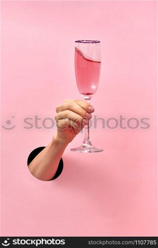 Woman hand from the hole holds glass with rose wine on a pastel pink background, copy space. Holiday concept.. Rose wine glass in a female&rsquo;s hand from the hole in the wall.