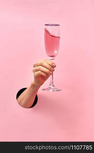 Woman hand from the hole holds glass with rose wine on a pastel pink background, copy space. Holiday concept.. Rose wine glass in a female&rsquo;s hand from the hole in the wall.