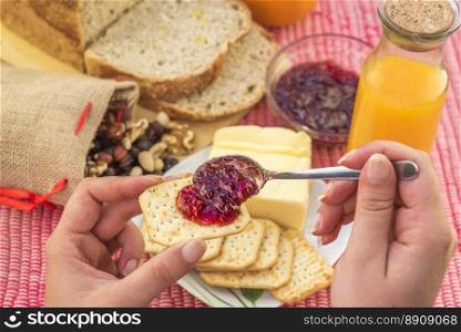 Woman hand holding a cracker with strawberry jam on it and in the background the breakfast table with bread, butter, orange juice and mixed nuts.
