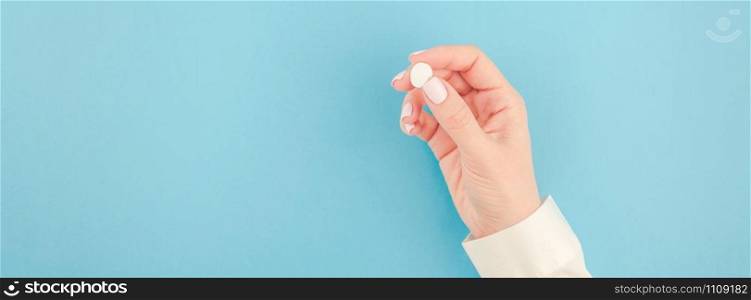 Woman hand holding a white pill on pastel blue paper background with copy space in minimal style, template for text. Concept of medical treatment, pharmacy, healthcare. Long wide banner