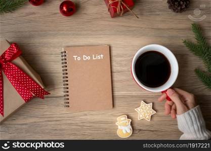 Woman hand holding black coffee cup with blank notebook and Christmas cookies on table. Xmas, Happy New Year, Goals, Resolution, To do list, Strategy and Plan concept