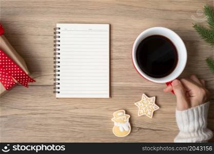 Woman hand holding black coffee cup with blank notebook and Christmas cookies on table. Xmas, Happy New Year, Goals, Resolution, To do list, Strategy and Plan concept