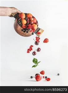 Woman hand holding copper cooking pot with falling summer fruits and berries  black and red currants, strawberries, raspberries, cherries, gooseberries and apricots and cherries at white background