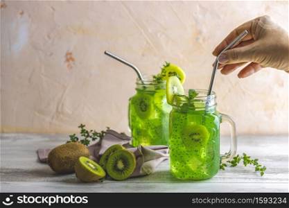 Woman hand holding drinking straw over glass jar of kiwi juice or smoothie. Kiwi Mojito cocktail or non-alcohol mocktail with mint and sliced kiwi fruits on wooden background copy space