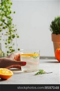 Woman hand holding glass with homemade lemonade with orange and thyme on grey concrete kitchen table. Refreshing summer drink with citrus fruit and herb. Front view.