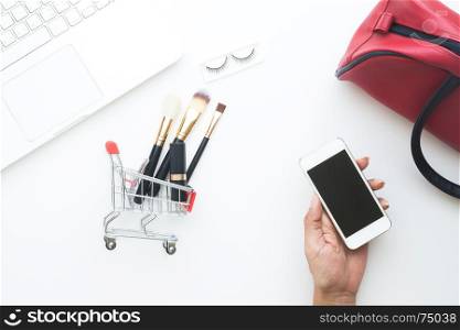 Woman hand holding mobile device with shopping cart and cosmetic items and laptop on white background, Top view online shopping concept
