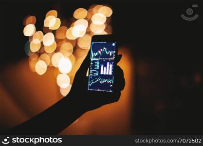 Woman hand holding mobile phone with stock trading on display in the dark with light bokeh blurry background, Financial, Commerce, Business and technology concept