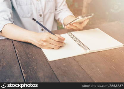 Woman hand holding phone and hand writing on table in garden at coffee shop with vintage toned.