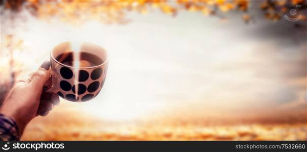 Woman hand holding polka dot cup with steamed hot coffee at autumn nature landscape with fall foliage and sunlight. Outdoor. Banner