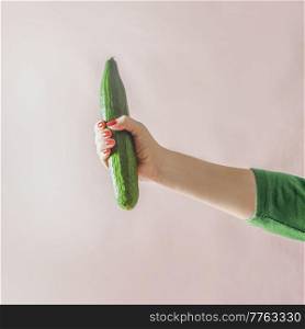 Woman hand holding whole raw green cucumber at pale beige wall background. Plastic free summer vegetable without packaging. Organic vegetable from garden. Front view.