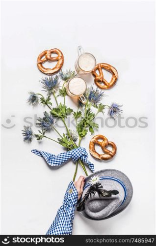 Woman hand in traditional blue shirt holding buch of alpine flowers on white background with pretzel and mugs of beer , top view. Oktoberfest concept. Modern layout. Flat lay