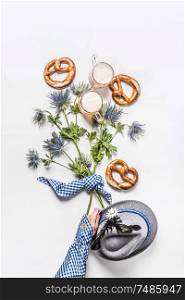 Woman hand in traditional blue shirt holding buch of alpine flowers on white background with pretzel and mugs of beer , top view. Oktoberfest concept. Modern layout. Flat lay