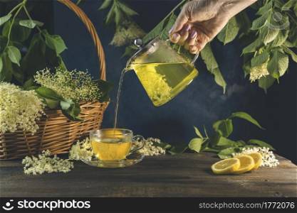 Woman hand is pouring hot healthy elder flowers tea from teapot to glass cup, dark surface