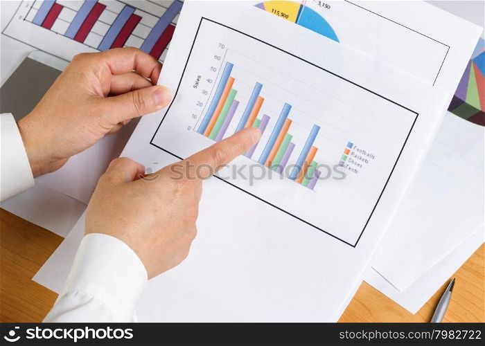 Woman hand pointing index finger at bar chart with printed graphs in background on desktop.