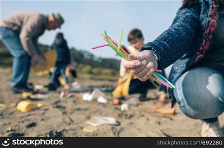 Woman hand showing handful of straws collected on the beach with group of volunteers working in the background. Selective focus in straws in foreground. Woman showing handful of straws collected on the beach