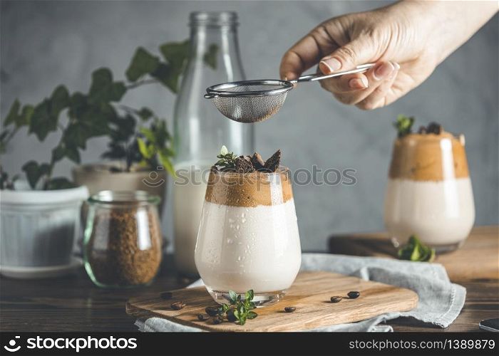 Woman hand sifting cocoa powder by sieve over glass of iced frothy drink Dalgona Coffee. Trend korean drink latte espresso with coffee. Dark rustic style.