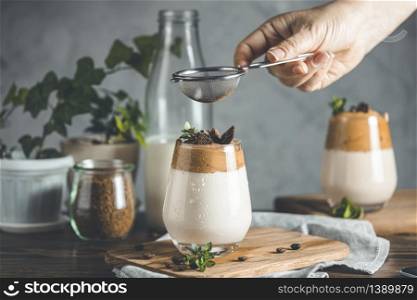 Woman hand sifting cocoa powder by sieve over glass of iced frothy drink Dalgona Coffee. Trend korean drink latte espresso with coffee. Dark rustic style.