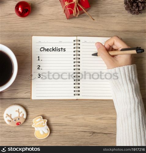 Woman hand writing checklist on notebook with black coffee cup and Christmas cookies on table. Xmas, Happy New Year, Goals, Resolution, To do list, Strategy and Plan concept