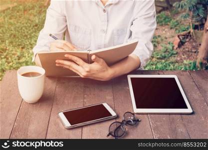 Woman hand writing notebook and phone, tablet on table in garden at coffee shop with vintage toned.