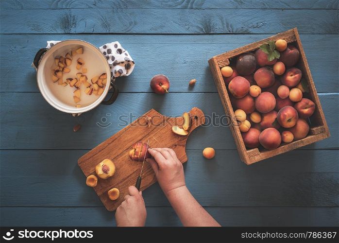Woman hands cutting into pieces peaches and apricots from a vintage wooden box, on a blue kitchen table. Above view with the making of peaches jam.