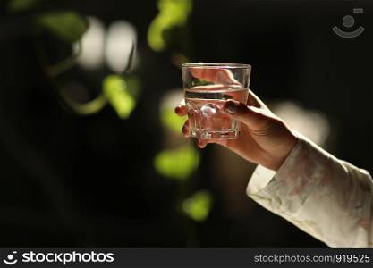 Woman hands isolated, holding a glass of water on a dark background with green leaves. the sun's rays fall on the glass.. Woman hands isolated, holding a glass of water on a dark background with green leaves. the sun's rays fall on the glass. healthy morning