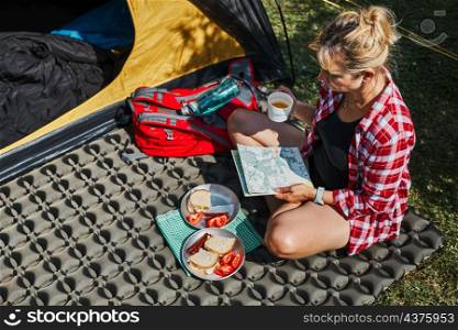 Woman having breakfast and planning day sitting by tent at camping. Female actively spending summer vacations close to nature outdoors. Concept of camp life