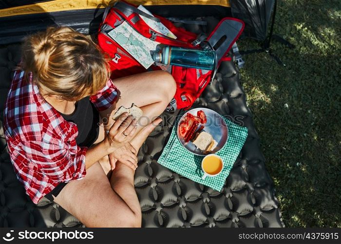 Woman having breakfast sitting by tent at camping. Female actively spending summer vacations close to nature outdoors. Concept of camp life