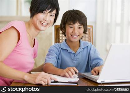 Woman helping young boy with laptop do homework in dining room smiling