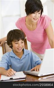 Woman helping young boy with laptop do homework in dining room smiling