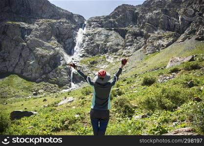 Woman hiker stands on the grass and enjoys waterfall view. Travel nature near beautiful waterfall Shaar in Kyrgyzstan. The highest waterfall in Central Asia.