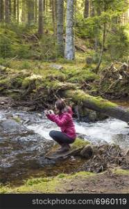 Woman hiker taking pictures. Young beautiful woman hiker taking pictures with its phone while sitting on a rock in the middle of a mountain stream in Black Forest, Germany