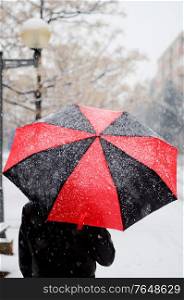 Woman holding a black and red umbrella under snow walking in a snowed street.. Woman holding a black and red umbrella under snow