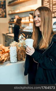 Woman holding coffee cup, standing in a coffee shop and waiting for the order. Smiling young woman having a break drinking coffee in cafe