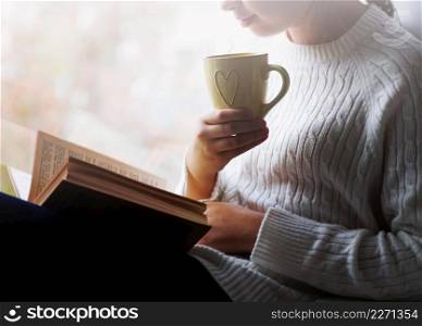 woman holding cup of tea and reading a book beside the window
