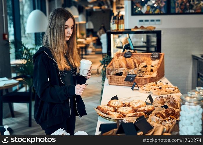 Woman holding cup with coffee looking at pastry, buns, cakes and cookies and waiting for the order. Girl buying a sweet food and hot drink to go. Young woman having a break doing shopping in a coffee shop