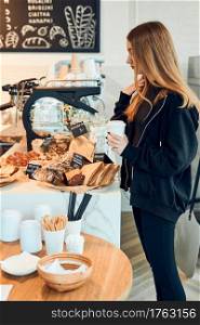 Woman holding cup with coffee looking at pastry, buns, cakes and cookies and waiting for the order. Girl buying a sweet food and hot drink to go. Young woman having a break doing shopping in a coffee shop
