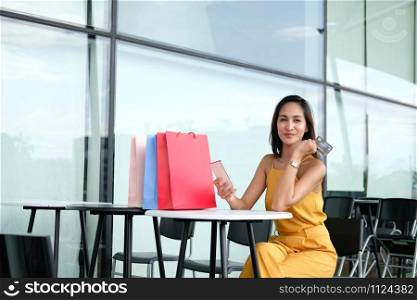 woman holding mobile smart phone & credit card for online payment with colorful shopping bags on table. consumerism lifestyle concept
