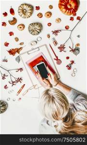 Woman holding smartphone with mock up screen on white desktop with notebooks, clipboard, fall leaves, pumpkins and cosmetics. Creative job layout. Home office. Top view. Blogging. Autumn flat lay