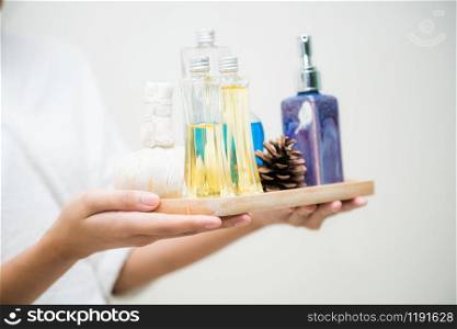 Woman holding spa treatment set including aromatic massage oil, body scrub lotion and herbal spa compress while wearing a bathroom suit.
