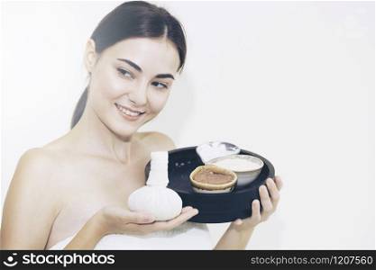 Woman holding spa treatment set including facial scrub lotion and herbal compress against white backgrounds. Luxury wellness and rejuvenation concept.