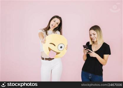 woman holding winking eye emoji near her friend using cellphone