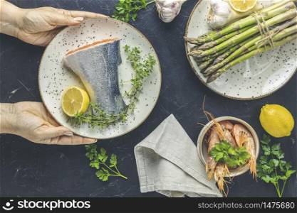 Woman holds ceramic plate with raw trout fish, thyme and lemon in hands on black concrete table surface surrounded plates with fresh raw asparagus, shrimp, prawn, parsley. Healthy seafood background.