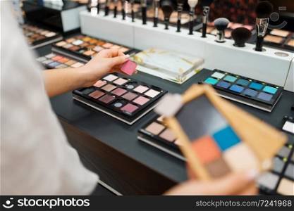 Woman holds eyeshadows at the shelf in cosmetics store. Buyer at the showcase in luxury beauty shop salon, female customer in fashion store. Woman holds eyeshadows at shelf in cosmetics store