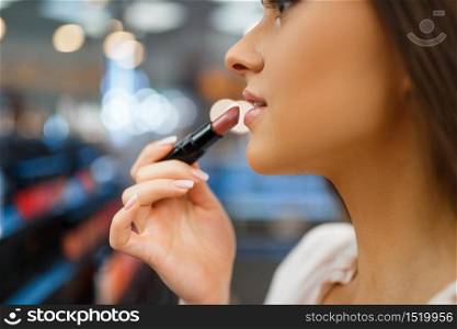 Woman holds lipstick at her face in cosmetics store. Buyer in luxury beauty shop salon, female customer in fashion market. Woman holds lipstick in cosmetics store