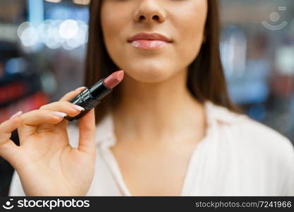 Woman holds lipstick at her face in cosmetics store. Buyer in luxury beauty shop salon, female customer in fashion market. Woman holds lipstick in cosmetics store