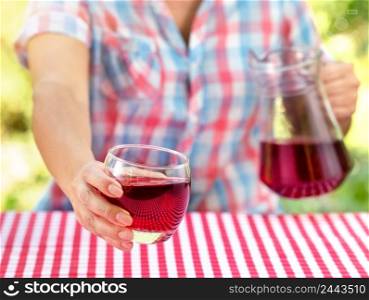 Woman holds out glass of wine or grape juice over table. Natural green background. Picnic concept. Woman holds out glass of wine or grape juice over table