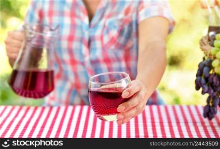 Woman holds out glass of wine or grape juice over table with red tablecloth. Nearby is a basket of grapes, in a hand a jug. Natural green background. Picnic concept. Woman holds out glass of wine or grape juice over table with red tablecloth